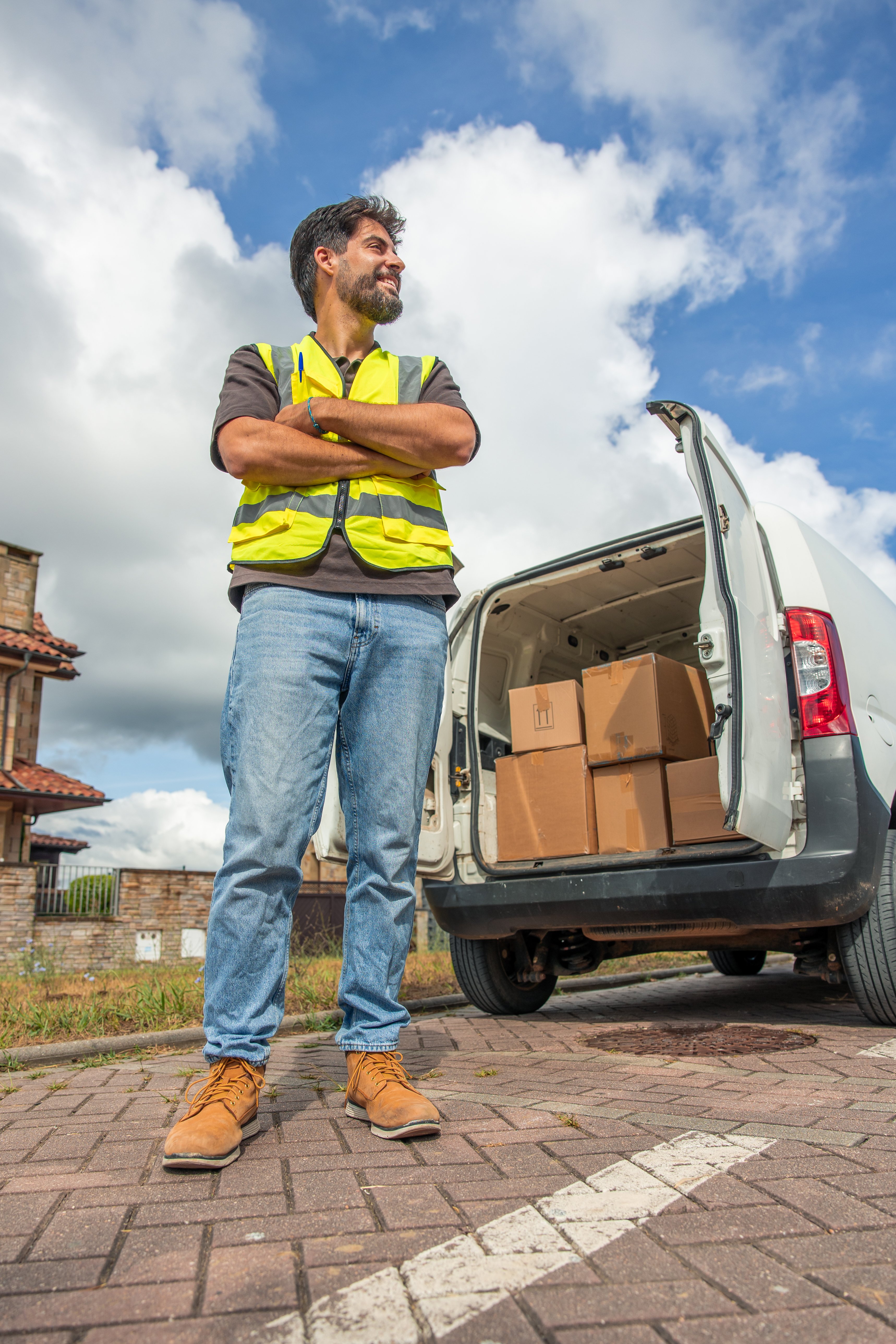 Smiling delivery driver wearing a high-visibility vest standing with arms crossed next to his cargo van's open trunk filled with cardboard boxes
